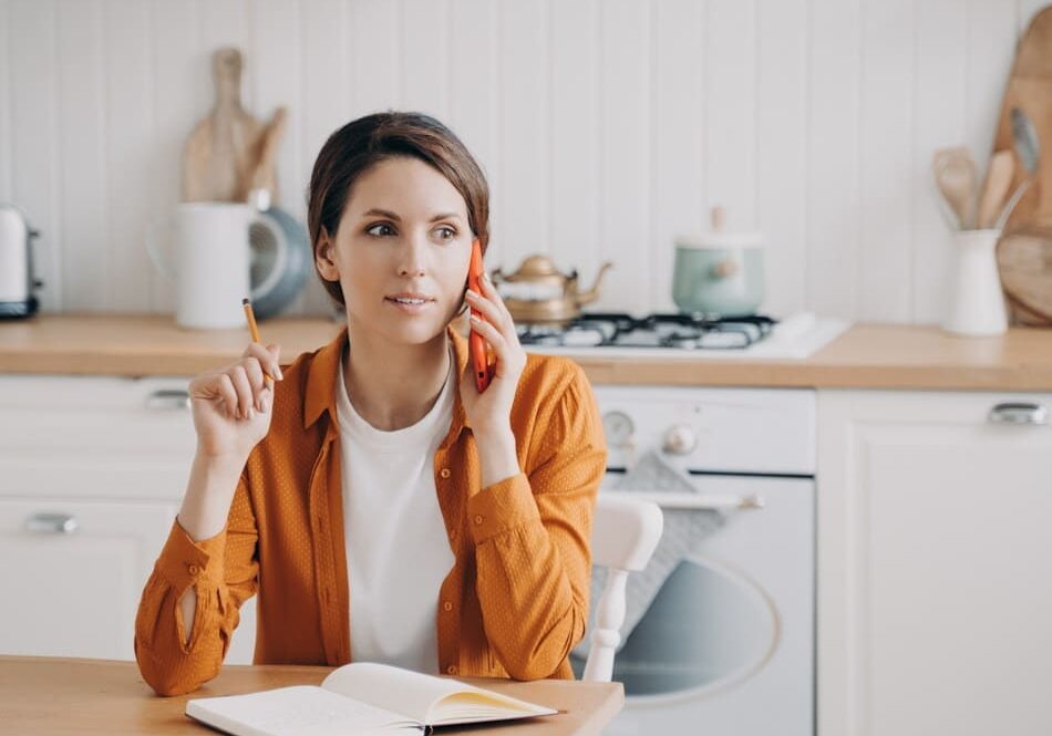 Busy female housewife talking on phone, calling to customer service, sitting in kitchen at home. Woman freelance worker discussing business issue, speaking on smartphone. Domestic life, remote job.